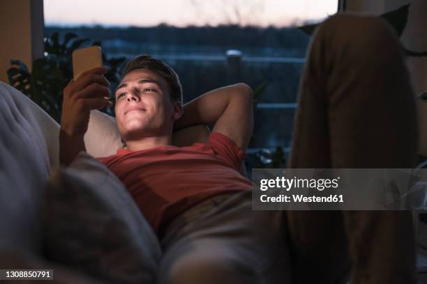 smiling young man lying on sofa while using smart phone in living room - 20 24 jahre stock-fotos und bilder