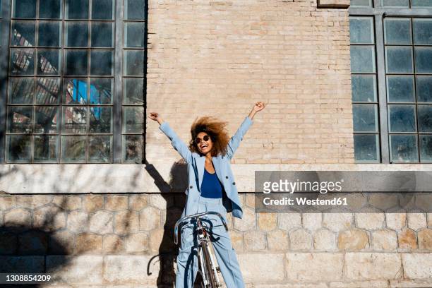 cheerful woman with arms raised standing with bicycle against building - green blazer stockfoto's en -beelden