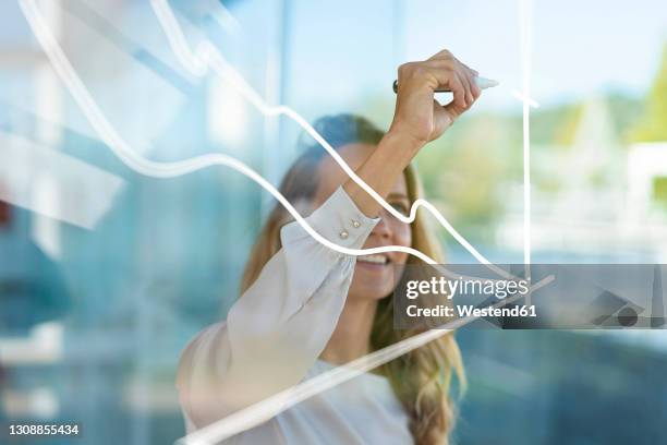 businesswoman drawing graph on glass while standing at modern office - diagramm stock-fotos und bilder