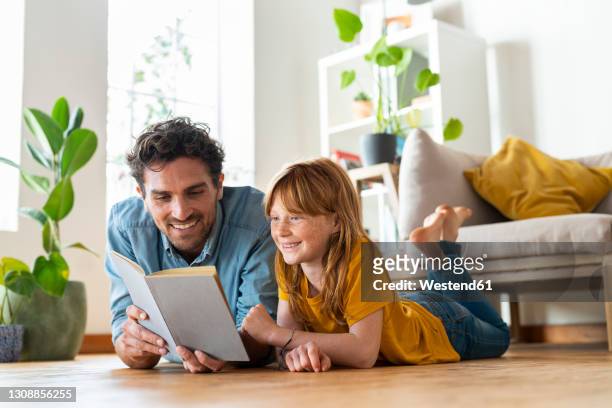 smiling father and daughter reading book together while lying down on floor at home - kid looking down stock pictures, royalty-free photos & images