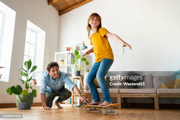 father helping daughter to ride skateboard in living room at home - skaten familie stock-fotos und bilder