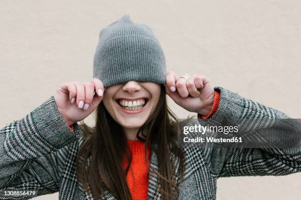 smiling woman covering eyes through knit hat against wall - knit hat stockfoto's en -beelden