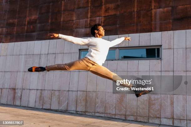 happy male dancer doing grand jet頡gainst building - mann freudensprung sonne vorderansicht leger stock-fotos und bilder