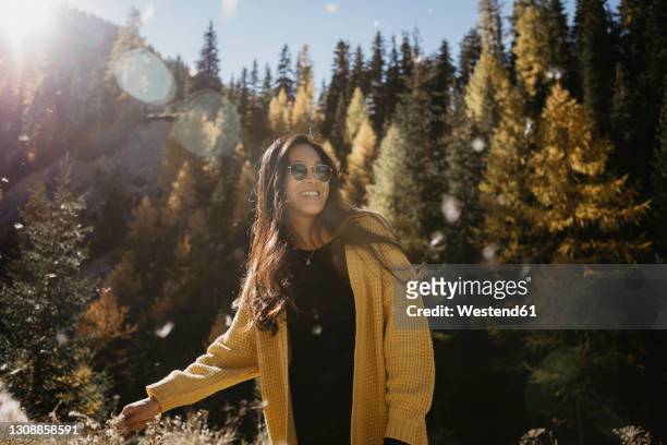 young woman wearing sunglasses smiling while standing in forest - smiling controluce foto e immagini stock