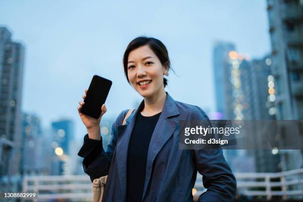 portrait of confident smiling young asian woman holding up her smartphone and standing against illuminated cityscape. mobile phone with blank screen for mobile app mockup design - happy corporate people with screen stock-fotos und bilder