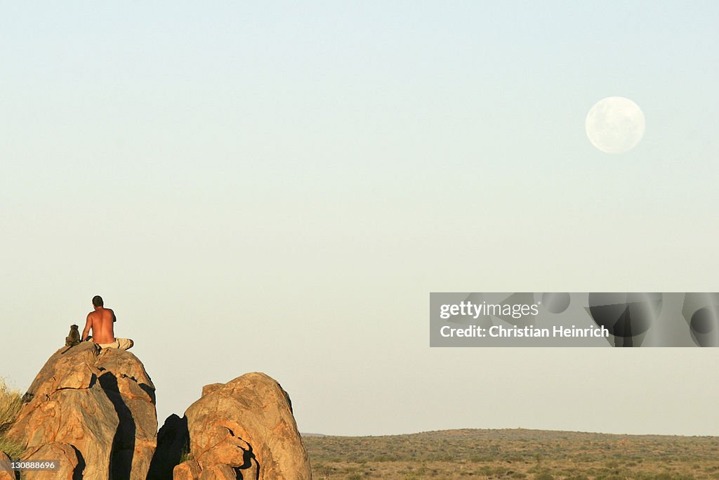 Namibian man sitting beside young tame Yellow Baboon (Papio cynocephalus) on a rock formation in late afternoon light watching the full moon rise over the grassland plains, Namibia, Africa