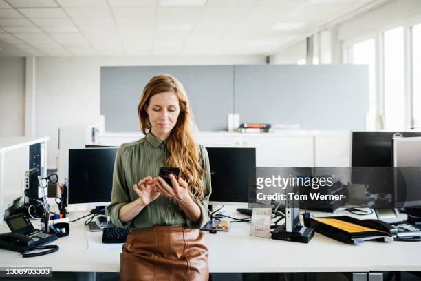 office manager perching on computer desk using smartphone - woman business desk front laptop office fotografías e imágenes de stock
