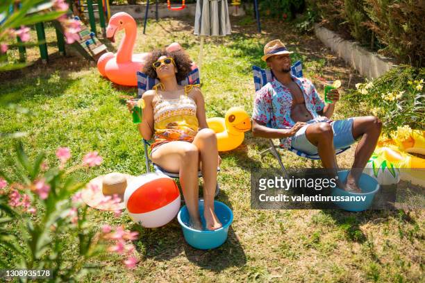 de paarzitting van afro op ligstoelen in de achtertuin en het hebben van zonnebaden tijdens hun staycation, tijdens pandemie - hot wife stockfoto's en -beelden