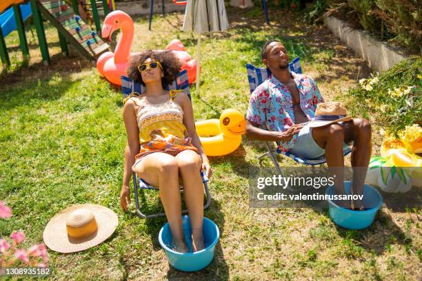 de paarzitting van afro op ligstoelen in de achtertuin en het hebben van zonnebaden tijdens hun staycation, tijdens pandemie - hot wife stockfoto's en -beelden