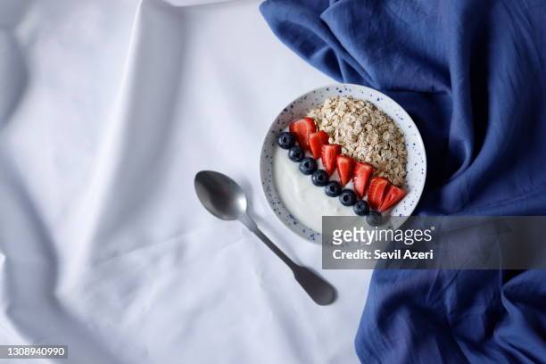 een kom gezond ontbijt dat met aardbeien en bosbessen op haveryoghurt op een witte dekking wordt verfraaid, naast het bord is een matte metaallepel en een blauw servet, het tendensontbijt van sociale media - blue bowl stockfoto's en -beelden