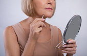 Cropped view of mature woman removing unwanted hair from her chin, using tweezers, looking in mirror on light background