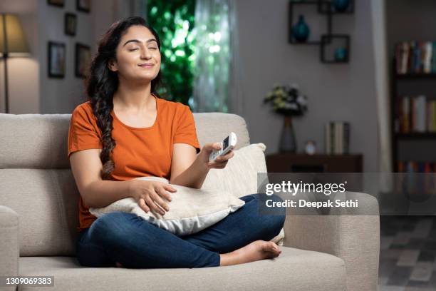 foto de una joven encendiendo el aire acondicionado sentado en el sofá en casa:- foto de archivo - refrescante fotografías e imágenes de stock