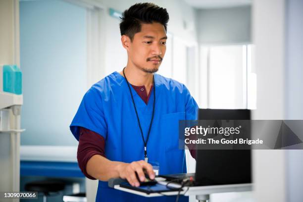 male nurse using laptop in hospital corridor - enfermeiro imagens e fotografias de stock