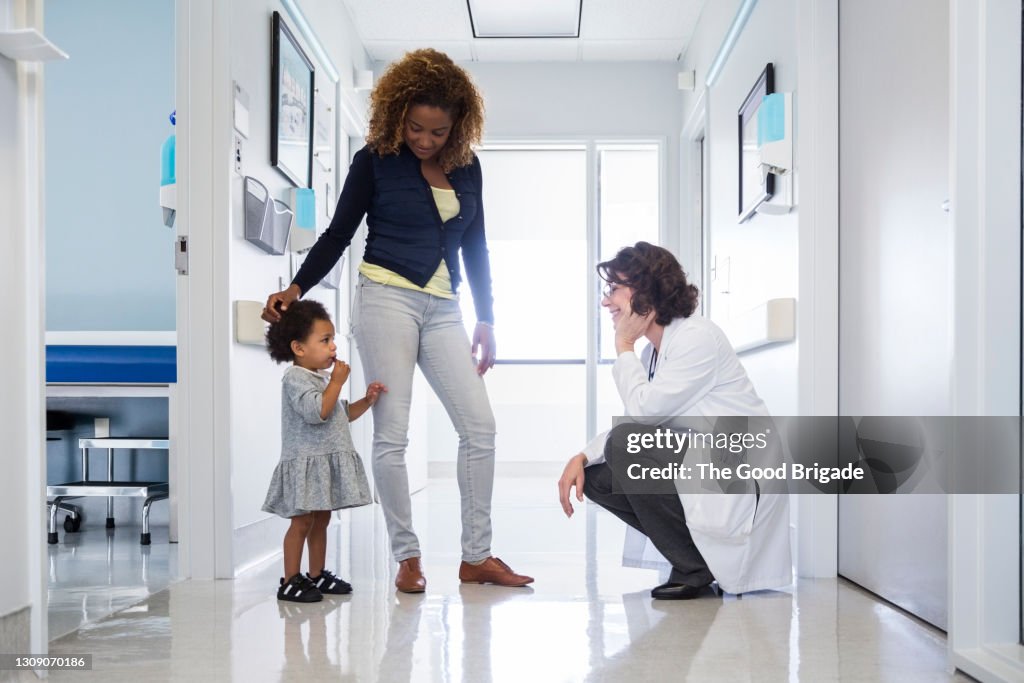 Smiling pediatrician talking to baby girl by mother in hospital corridor