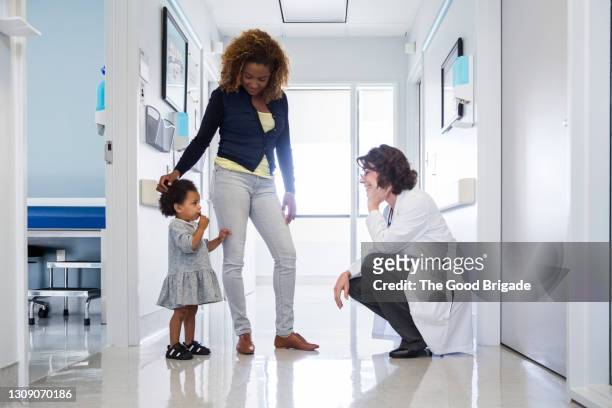 Smiling pediatrician talking to baby girl by mother in hospital corridor