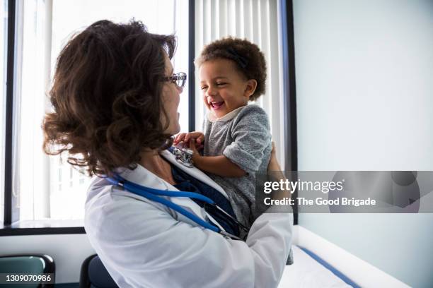 female doctor holding cheerful baby girl in medical exam room - pédiatre photos et images de collection