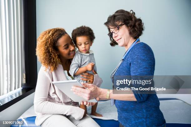 female pediatrician showing digital tablet to mother while holding toddler in hospital - medical examination of young foto e immagini stock