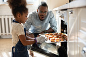 Excited african dad watching daughter taking muffins out of oven
