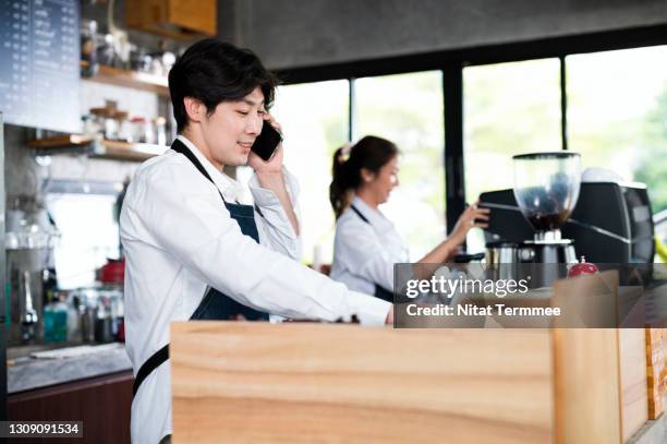 smiling asian male owner taking order with customer on a cellphone in a coffee shop. food and beverage ordering system, customer service mind and small business owner. - busy coffee shop stockfoto's en -beelden