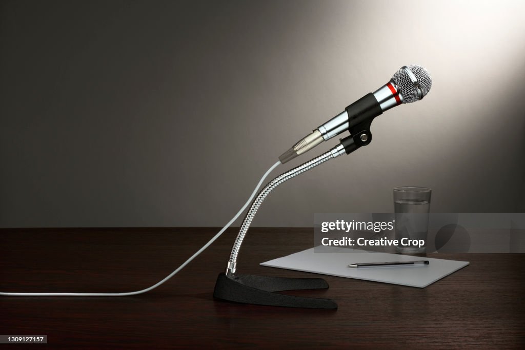 A microphone in a stand, with paper and a glass of water, all on a wooden desktop