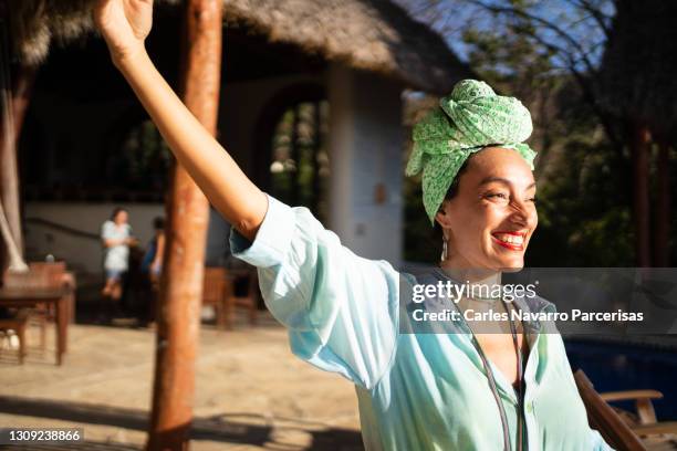 smiling woman in a caribbean dress raising the arm in front of a swimming pool with the sun illuminating her face - laufsteg fashion stock-fotos und bilder