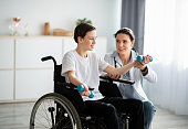 Physical rehabilitation concept. Young physiotherapist helping teenage boy in wheelchair to do exercises at home