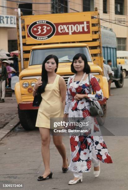 Two young Vietnamese women, one dressed in a pale yellow minidress, with the other wearing a floral print Ao Dai, walking hand-in-hand, with a '33...