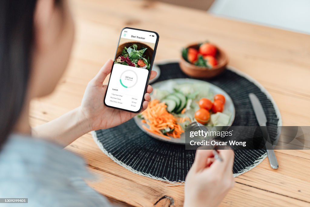 Young Woman Using Calorie Counter App On Smartphone While Eating Salad