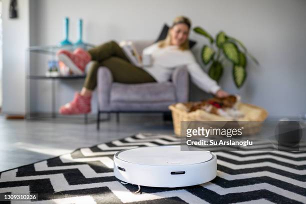 young woman petting her dog while robot vacuum cleaner is cleaning the house - robot vacuum stock pictures, royalty-free photos & images