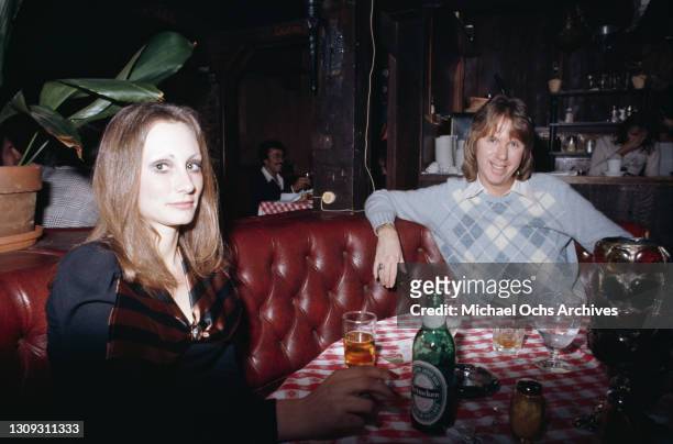 Couple sitting on a button back bench seat at a table, the woman holds a lit cigarette, with a bottle of Heineken on the table before her, with the...