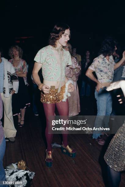 Male clubber wearing a green cropped floral print, short-sleeved blouse with a gold sequin miniskirt and red tights among the dancers on the...