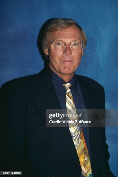 American actor Adam West wearing a dark blue blazer over a blue shirt with a patterned tie, against a blue background, location unspecified, 1993.