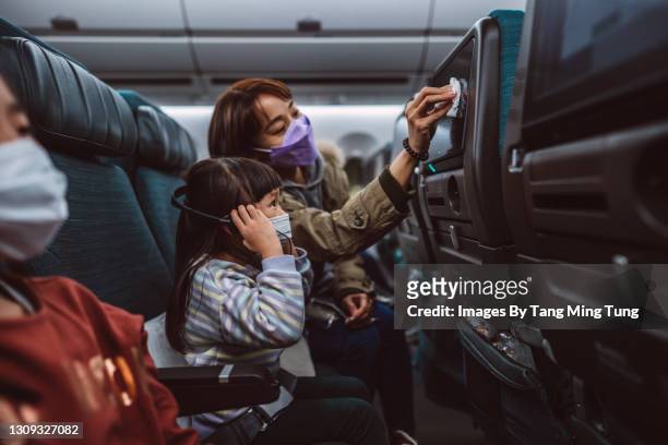 Mom & daughter in protective face masks cleaning the seat-back TV screen with disinfectant surface wipe while travelling on the airplane