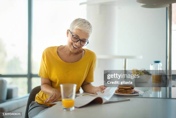 mujer madura leyendo una revista mientras desayunaba. - mujer revista fotografías e imágenes de stock