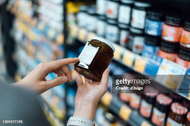 close up of young woman grocery shopping in a supermarket. standing by the aisle, holding a bottle of marmalade, reading the nutritional label and checking ingredients at the back - information nutritionnelle photos et images de collection