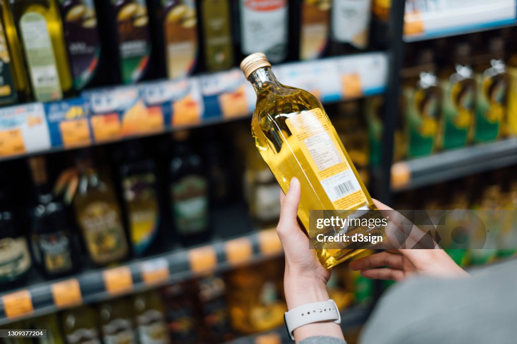 Close up of young woman grocery shopping in a supermarket. Standing by the aisle, holding a bottle of organic cooking oil, reading the nutritional label and checking ingredients at the back