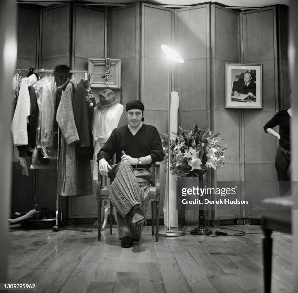 British fashion designer John Galliano wearing baggy striped trousers, a black bandana head covering and a V-neck sweater is pictured seated smoking...