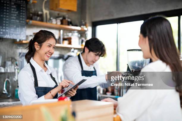 a young asian women waiters are taking orders from customer while working behind cashier counter in a small coffee shop. food and beverage ordering system and small business concepts. - coffee shop owner stockfoto's en -beelden