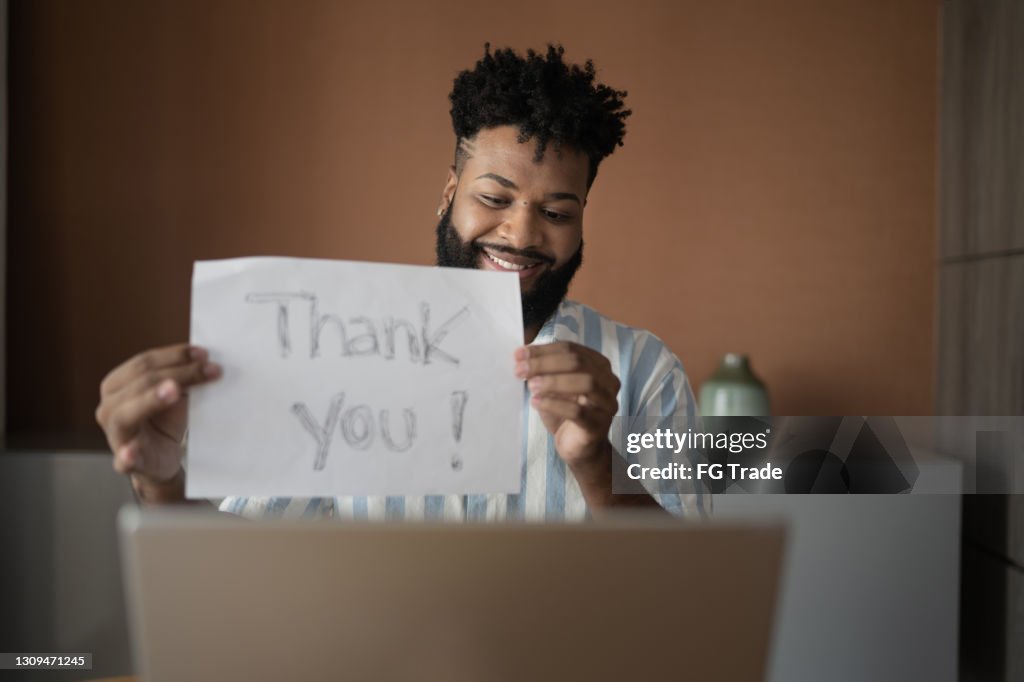 Junger Mann hält Papier mit "Dankeschön"-Text auf einem Videoanruf am Laptop zu Hause