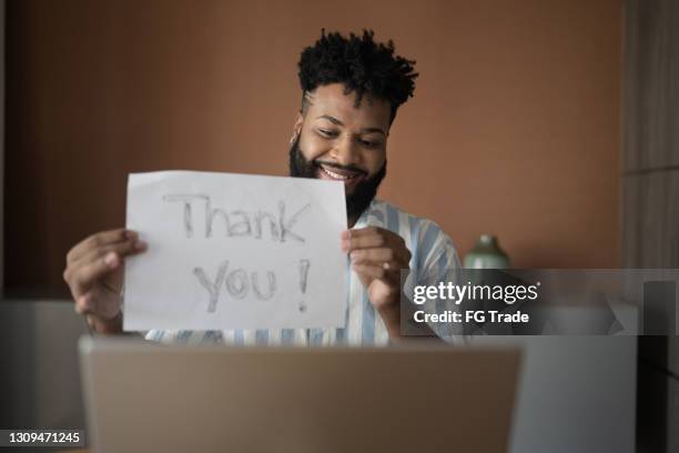 junger mann hält papier mit "dankeschön"-text auf einem videoanruf am laptop zu hause - thank-you-englischer-satz stock-fotos und bilder