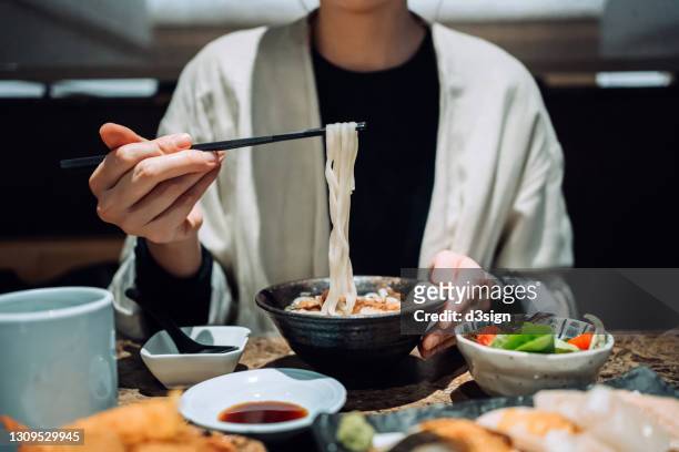 close up mid-section of young asian woman eating a bowl of japanese udon soup noodles, together with assorted sushi, tempura and vegetable salad freshly served on the dining table in a restaurant. asian cuisine and food. eating out lifestyle - udon noodle stock pictures, royalty-free photos & images