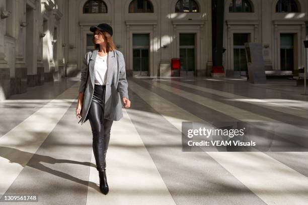 stylish woman wearing white t-shirt, long blazer, black leather pants and black cap walking alone through a public corridor. - high fashion stock-fotos und bilder