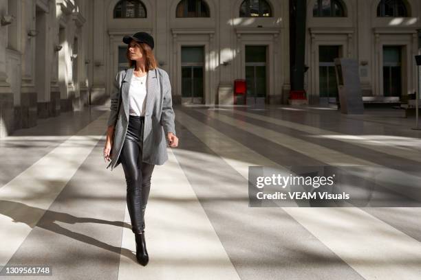 stylish woman wearing white t-shirt, long blazer, black leather pants and black cap walking alone through a public corridor. - chic fashion stock pictures, royalty-free photos & images