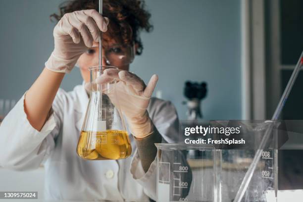 giovane scienziata che indossa dispositivi di protezione prendendo un campione di liquido giallo da una fiaschetta erlenmeyer - pallone di vetro foto e immagini stock