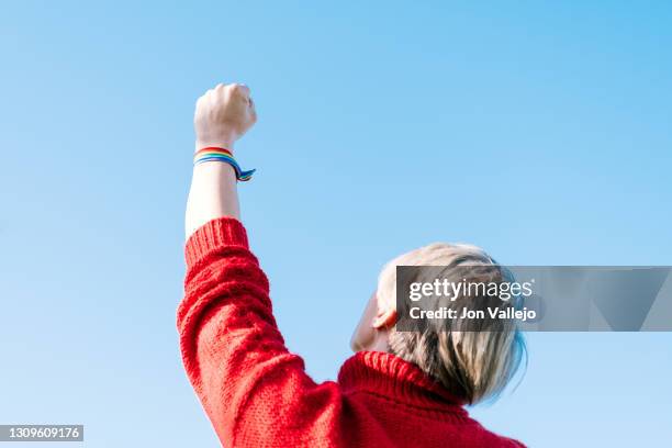 a lesbian woman in a red sweater is holding her fist up in a sign of struggle. she is wearing a pride bracelet. - militant photos et images de collection