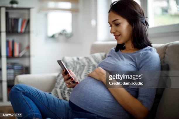 smiling pregnant woman using mobile phone at home - pregnant lady stock pictures, royalty-free photos & images