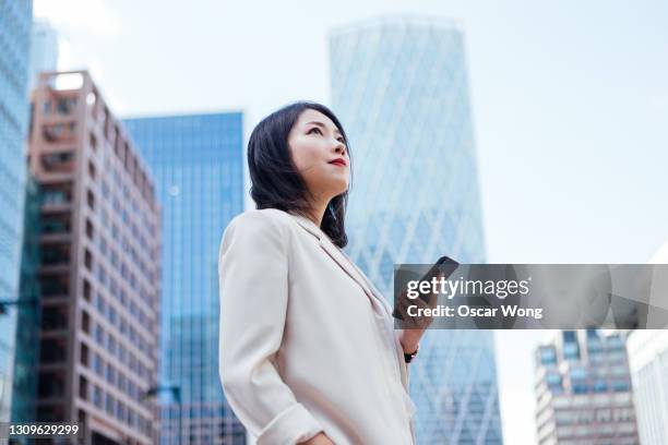 confident young businesswoman with smartphone standing against modern buildings - japanese woman looking up stock pictures, royalty-free photos & images