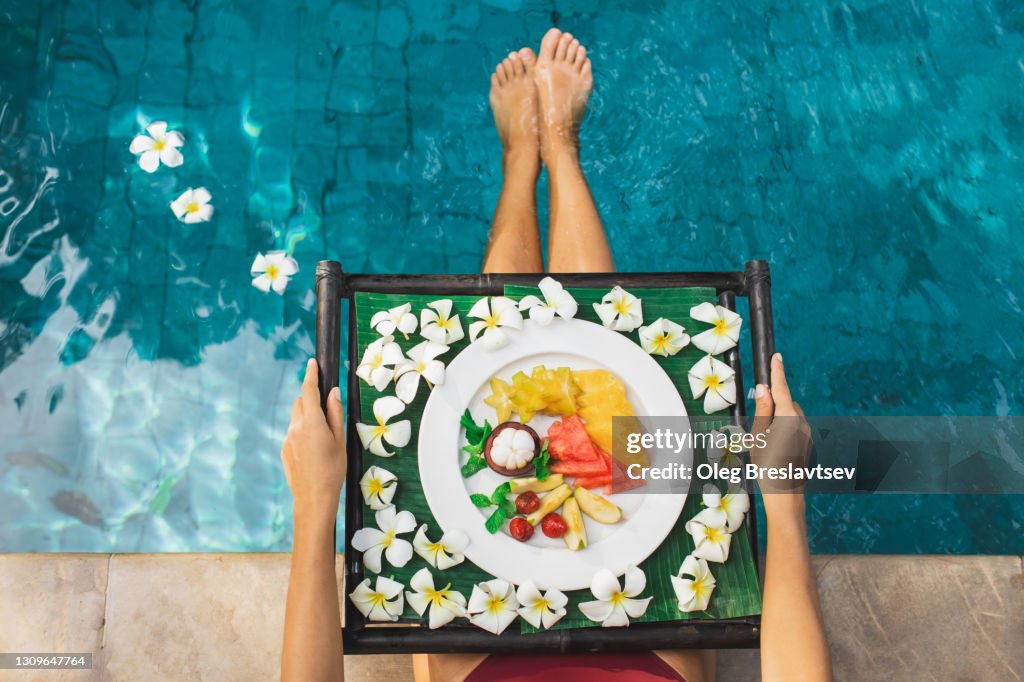 Woman sitting on poolside and holding tray with fresh tropical fruit breakfast. Unrecognizable person