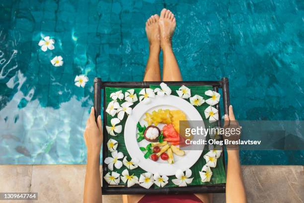 woman sitting on poolside and holding tray with fresh tropical fruit breakfast. unrecognizable person - holiday healthy eating stock pictures, royalty-free photos & images
