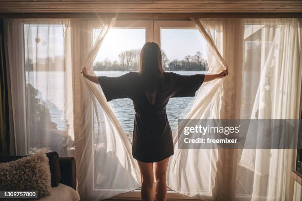 back view of woman in bathrobe opening curtains and looking out - window curtains stock pictures, royalty-free photos & images